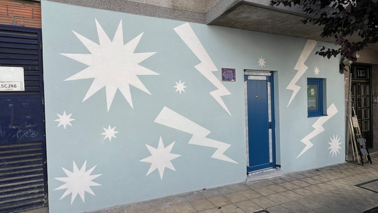 Mural en la fachada de una vivienda: estrellas y rayos blancos sobre un fondo azul claro, alrededor de una puerta y una ventana azules