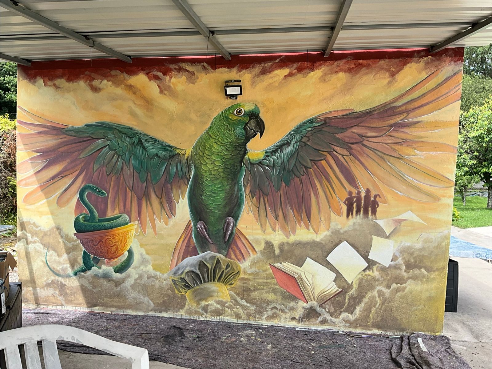 Tarzán: mural de un loro verde con las alas abiertas sobre un mar de nubes, con una serpiente enroscada en una copa, un libro abierto y cuatro siluetas